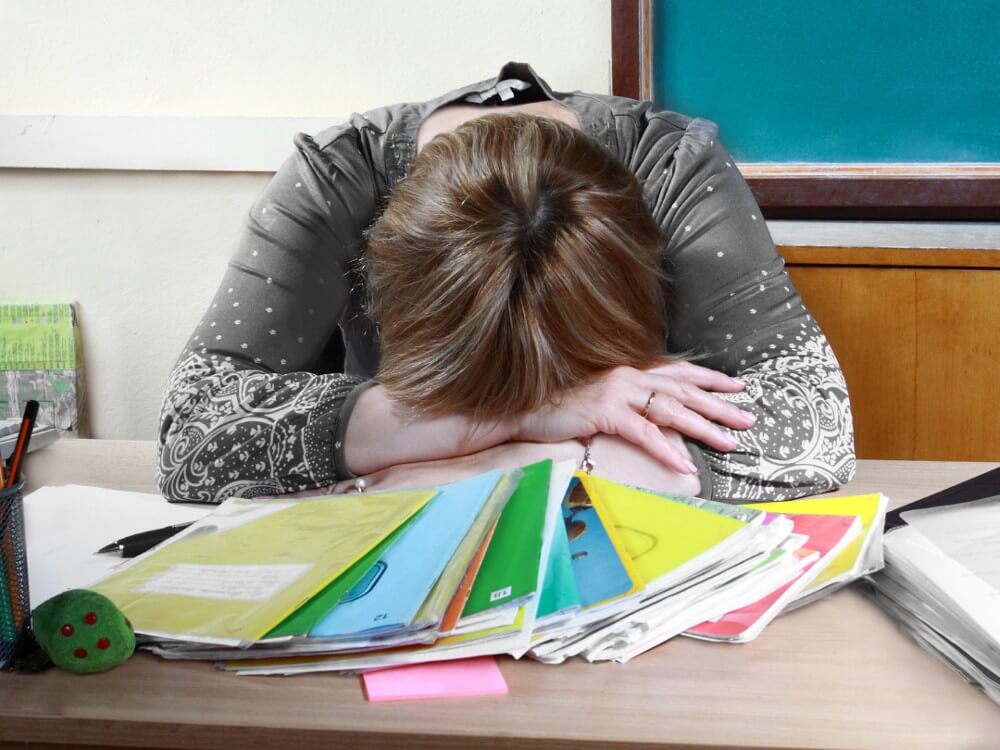 An unhappy teacher rests her head on the desk defeated by her lack of work-life balance.