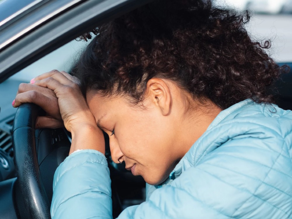 An exhausted woman experiencing teacher burnout with her head against a steering wheel.