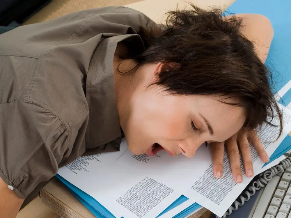 A female teacher yawning while laying her head on a desk, exhausted from teaching.