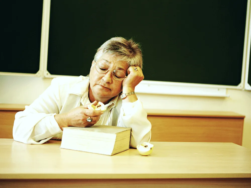 A tired teacher asleep at her desk while eating an apple.