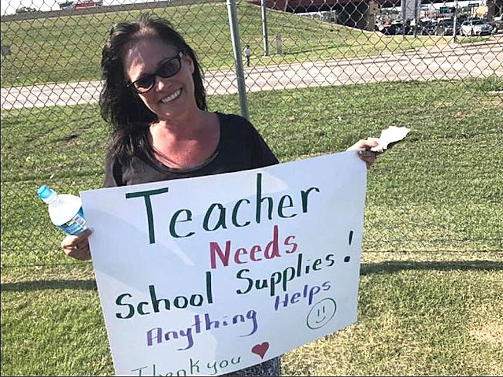 Teacher holding a sign that says, "Teacher needs school supplies! Anything helps.'