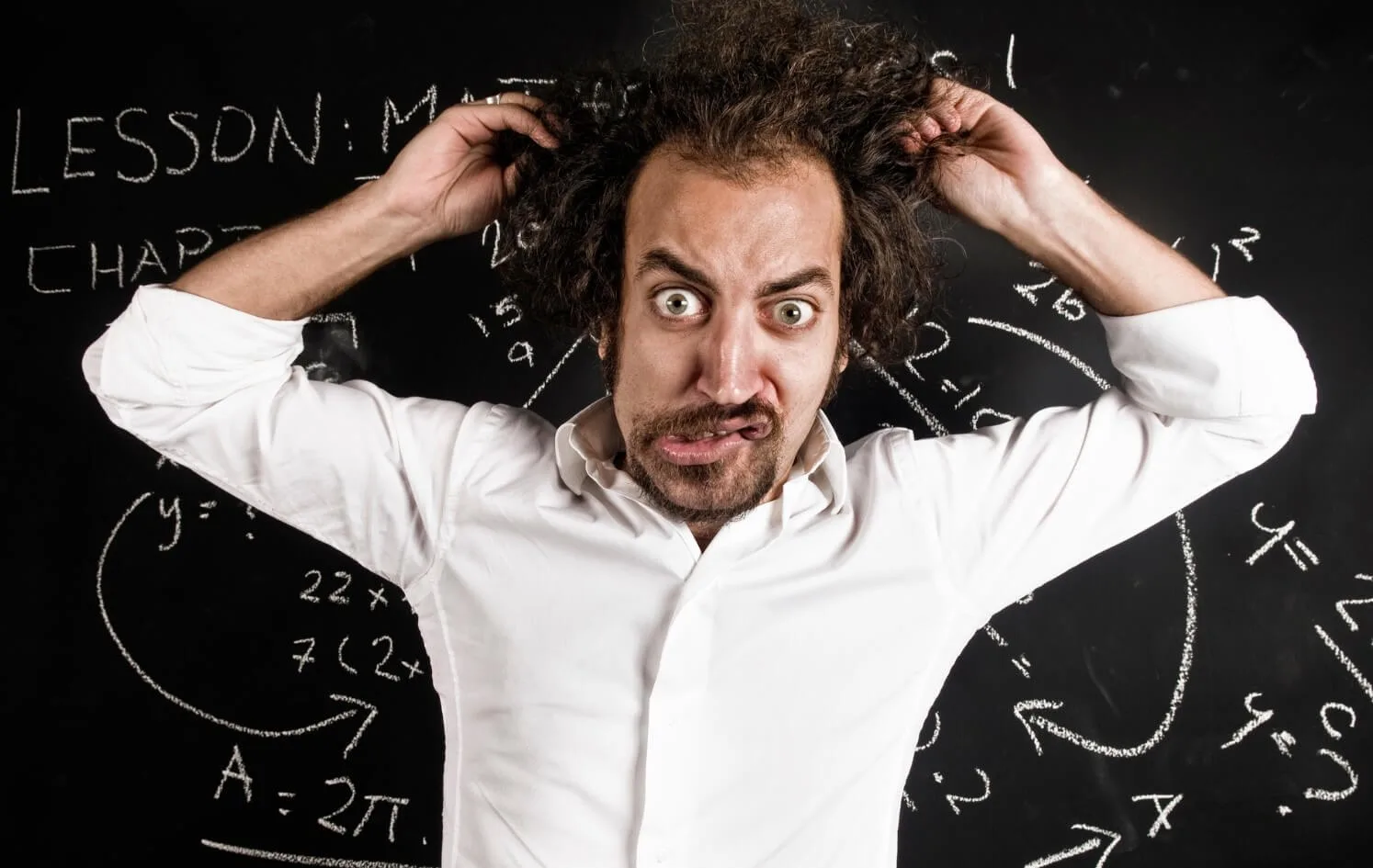 A man who hates his teaching job pulling his hair out in front of the classroom blackboard.