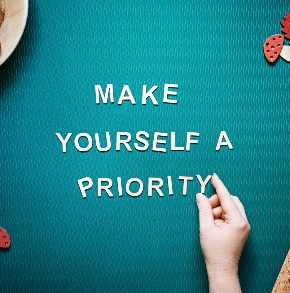 A hand placing letters on a blue table spelling out "Make Yourself a Priority" in reference to the list of self-care teacher burnout recovery strategies.